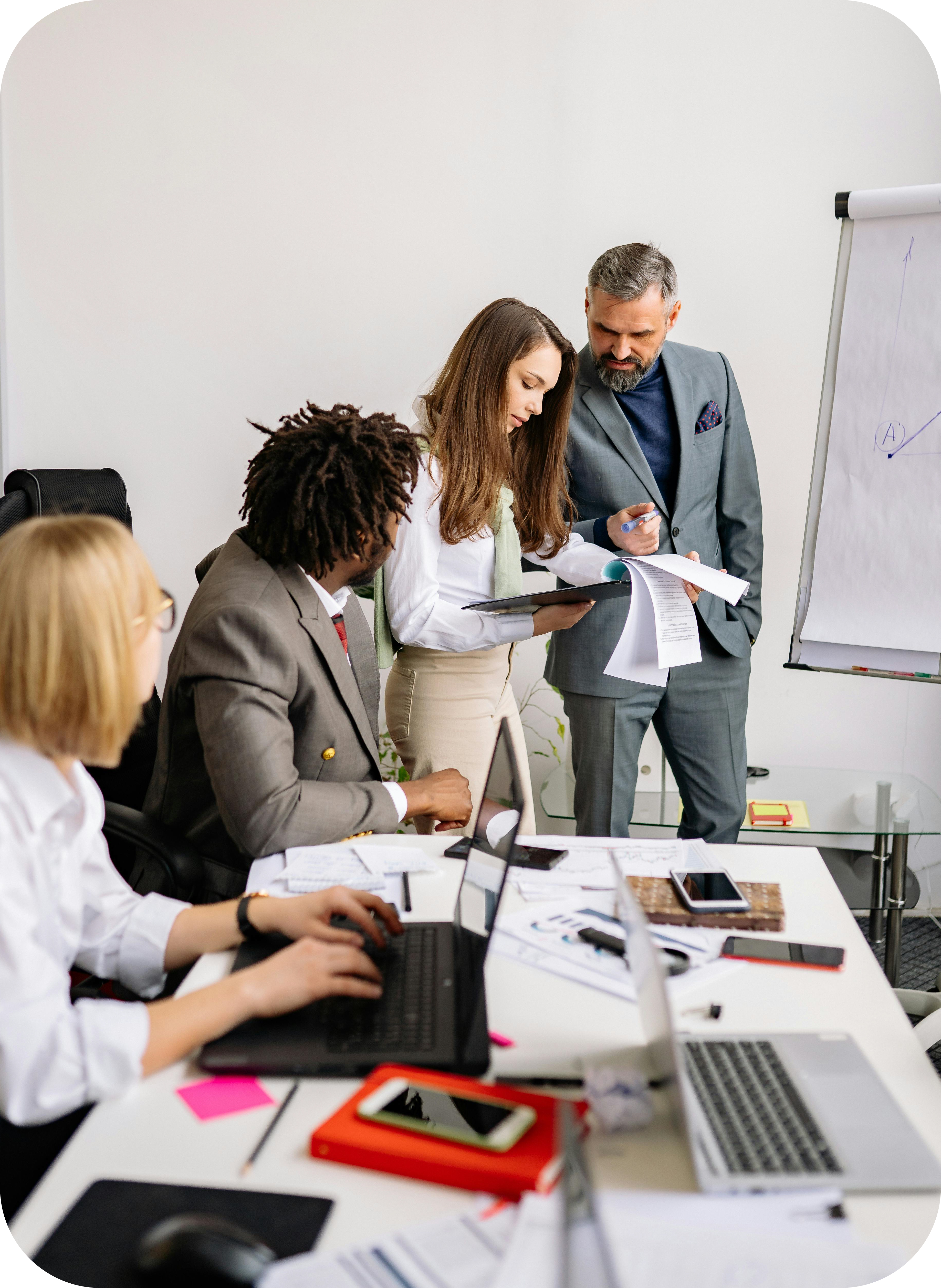 Image of 4 people (two standing and two sitting at the table in the foreground) in an conference room discussing a document that the two standing people are holding and looking at.